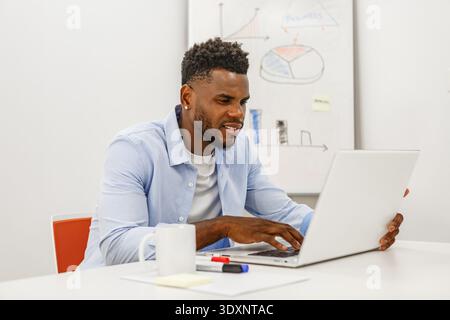 A focused young man sits at a desk, typing on his laptop with a mug and markers nearby. He is engaged in some work amidst charts on the whiteboard beh Stock Photo
