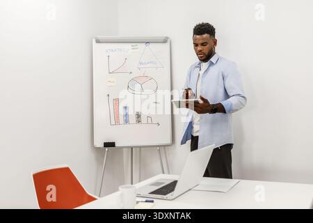 A businessman stands next to a whiteboard displaying various charts and graphs while using a tablet to present data. A laptop and coffee cup are on th Stock Photo