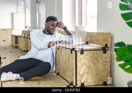 A person is seated on wooden boxes, focused on a laptop in a bright and modern coworking environment. The workspace features large windows and a conte Stock Photo