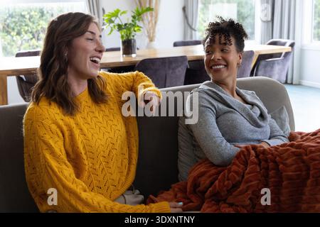 Diverse female friends wearing mustard, gray knits talking on gray sofa at home with rust throw Stock Photo