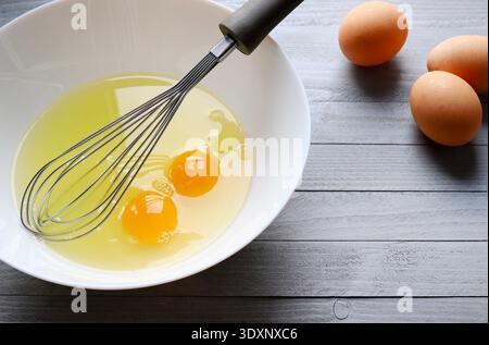 A white bowl with two cracked eggs and a hand whisk on a gray wooden background. Concept of culinary creation. Stock Photo