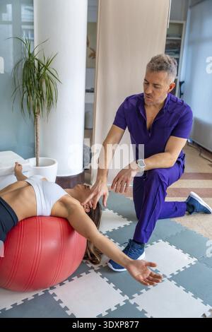 Male physiotherapist assisting a young woman client stretching and doing an exercise on a red stability ball in a professional medical recovery, rehab Stock Photo