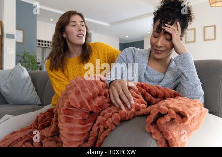 Diverse female friends sitting on gray sofa at home, comforting under rust-orange throw blanket Stock Photo