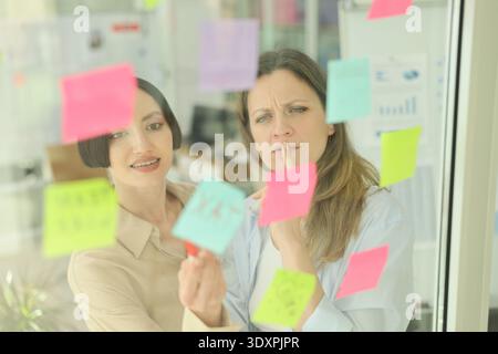 Two women collaborating at a bright office space while brainstorming ideas on colorful sticky notes during a busy workday Stock Photo