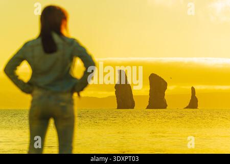Out of focus woman watches dramatic sea stacks silhouetted by a golden yellow sunset in the ocean on the horizon. Dreamy view from the coast evokes an Stock Photo