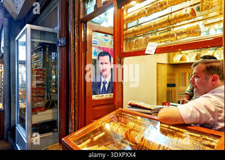 People explore Al-Hamidiyah Souq in Damascus, browsing shops for goods. A large portrait of former president Assad hangs on the wall next to a vendor. Stock Photo