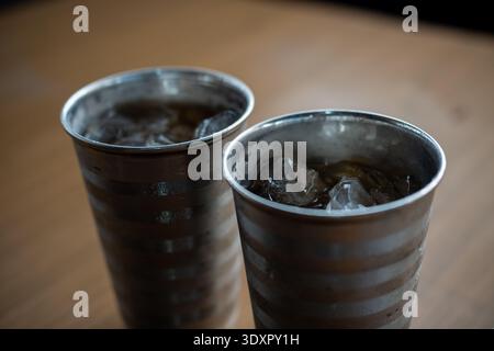 Two metal cups filled with dark liquid and ice cubes on wooden table Stock Photo