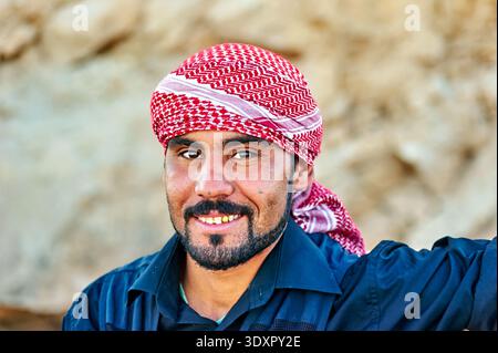 A young man stands outdoors in Syria, wearing a red and white headscarf. He smiles and looks at the camera with a friendly expression. Stock Photo