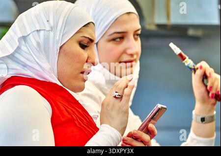 Two young women share a moment in Aleppo, Syria, smoking hookah and using their mobile phones while enjoying each other's company. Stock Photo