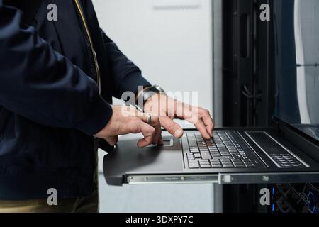 IT technician typing on a laptop in a server rack in a data center performing network maintenance Stock Photo