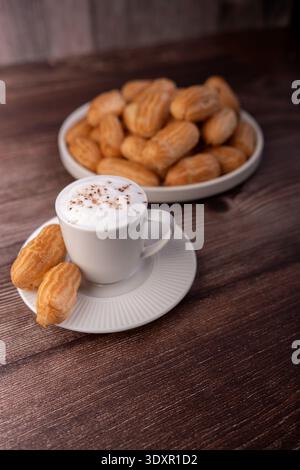 close-up of a frothy cappuccino in a white ceramic cup and saucer with two golden eclairs beside it, a bowl of pastries in the blurred background on a Stock Photo