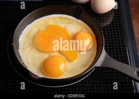 close-up overhead photograph of three cracked eggs in a small black cast-iron skillet on a dotted glass induction cooktop, vivid golden-orange yolks g Stock Photo