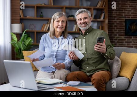 Happy senior couple sitting on a sofa, holding bills and a smartphone while using a laptop to review and manage expenses, representing financial planning and successful budgeting at home Stock Photo