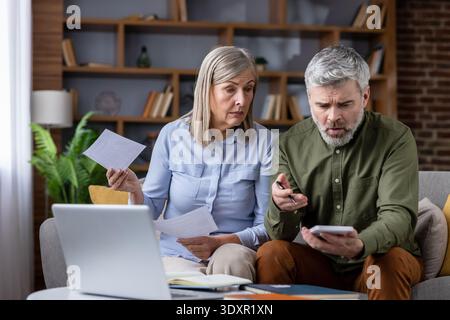 Senior couple sitting on a couch, focused on paperwork and a calculator, discussing bills and tax problems, experiencing worry and financial pressure at home Stock Photo