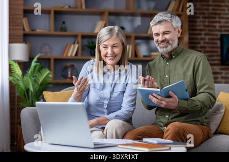 Senior couple on a couch having a cheerful video conference using a laptop, discussing retirement plans and financial strategy with a professional from their home office environment Stock Photo