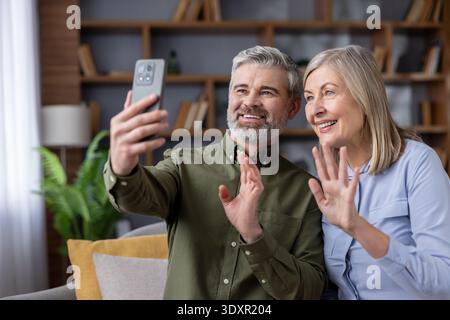 Happy senior couple sitting on a couch, smiling and waving to a handheld smartphone during a video call, communicating with family or friends via technology at home Stock Photo