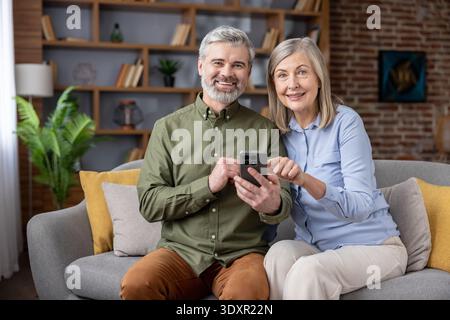 Senior couple sitting on a sofa, happily using a smartphone together, enjoying modern technology for communication and connection in their comfortable living room Stock Photo