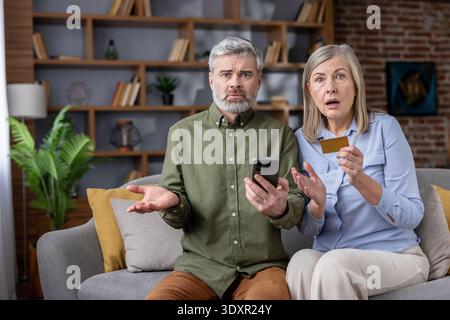 Shocked senior couple sitting on a couch, looking at a smartphone and holding a credit card, feeling confused and worried about a potential online banking scam or financial loss Stock Photo
