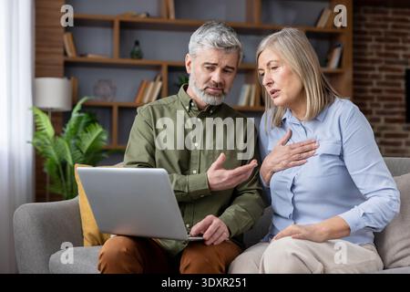 Mature couple on sofa, worriedly reading laptop, woman clutching chest with shortness of breath while man gestures in concern, discussing health, finances and bad news Stock Photo