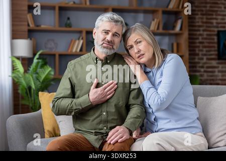 Mature man suffering from chest pain, holding his hand to his heart while his concerned wife comforts him at home, representing health issues, medical emergencies, and the stress of aging Stock Photo