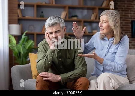 Senior couple sitting on a sofa, woman talking loudly and gesticulating, man looking sad and frustrated, facing relationship problems and marital conflict in their home Stock Photo