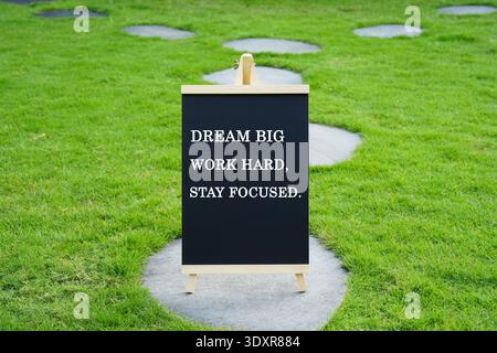 A black chalkboard on an easel in a green garden featuring the motivational quote "DREAM BIG, WORK HARD, STAY FOCUSED." Stock Photo