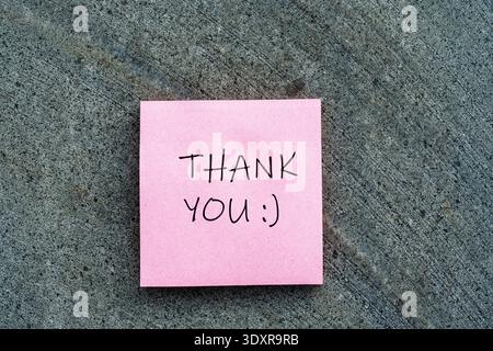 Pink sticky note with "THANK YOU :)" handwritten text on a grey stone texture, representing gratitude, appreciation, and positive feedback Stock Photo