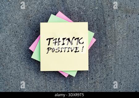 "THINK POSITIVE!" handwritten on a stack of colorful sticky notes over a grey stone background. Stock Photo
