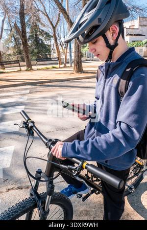 Young male cyclist wearing helmet and backpack, checking directions on his smartphone while standing next to bike Stock Photo