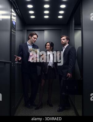 Three young professionals standing in office elevator using smartphones and holding flowers Stock Photo