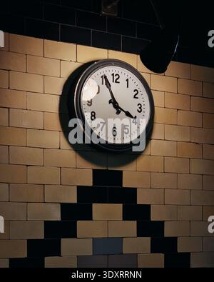 Old clock on beige tiled wall in a dark room. Old analog railway clock with white clock face and thick black bezel mounted on beige brick wall. Stock Photo