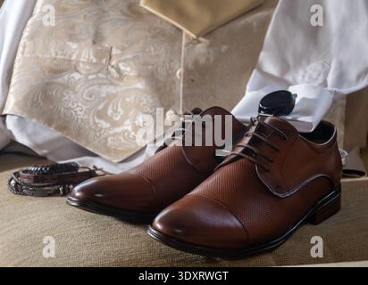 Elegant brown leather dress shoes placed beside groom accessories including a patterned beige waistcoat, tie, cufflinks, belt and wristwatch. Classic. Stock Photo
