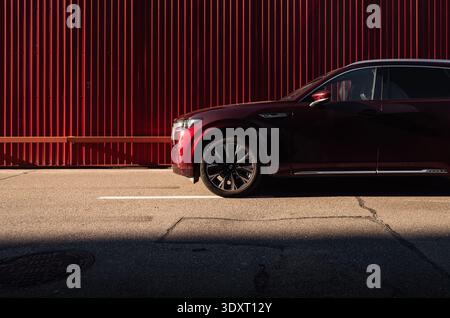 Dark red SUV on the street in industrial district. Front side part view of Mazda CX-90 7-seater parked against red corrugated wall on sunny day. Stock Photo