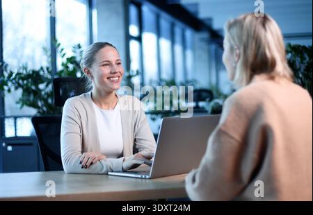 Two women are sitting at a desk with a laptop in front of them Stock Photo