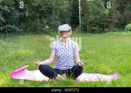 A woman is in a meditation pose, using virtual reality goggles for therapy and cognitive rehabilitation. An innovative blending of nature and digital Stock Photo