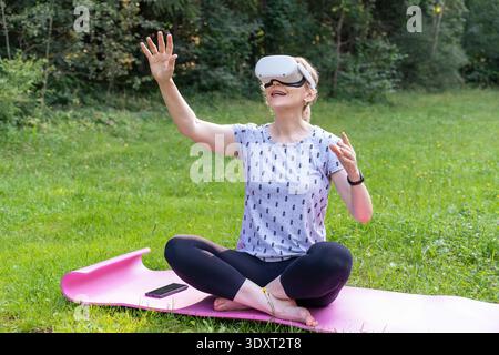 An excited woman interacts with a virtual gaming world while sitting on a yoga mat in nature. Stock Photo