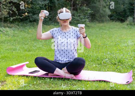 A woman performs therapeutic exercises using VR goggles and controllers while sitting on a yoga mat. Stock Photo
