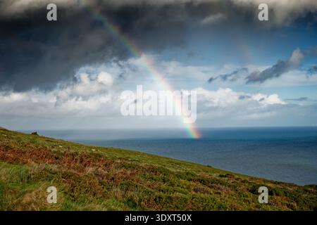 The Sun breaks though dark rain clouds over the sea with a bright rainbow plunging into the water, Great Orme, Llandudno, North Wales. Stock Photo