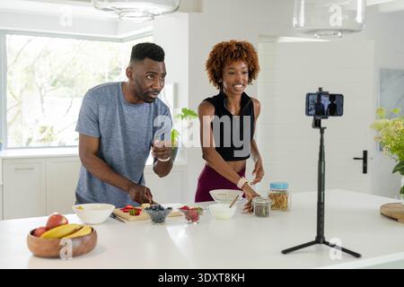 Diverse couple preparing food at kitchen island, recording on smartphone tripod with cutting board Stock Photo