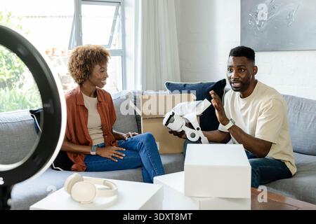 Diverse couple sitting on gray sofa at home holding white VR headset and smiling Stock Photo