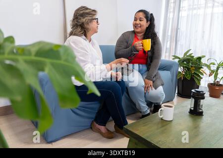 Two women relaxing on a couch, drinking coffee and talking together in a comfortable home Stock Photo