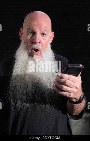 Portrait of old bald man with a long white beard using phone against a dark background Stock Photo
