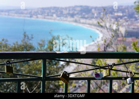 Colorful love locks including heart-shaped ones attached to a green railing with breathtaking panoramic view of Nice's turquoise Baie des Anges below. Stock Photo