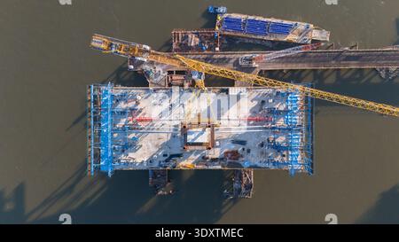 Aerial view of a massive bridge section under construction with cranes and equipment casting long shadows on the water, Novi Sad, Vojvodina, Serbia. Stock Photo