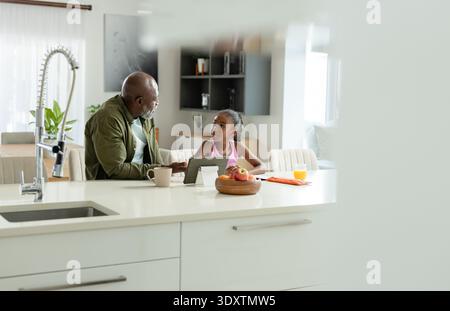 African American father and child talking at kitchen island using tablet holding coffee mug Stock Photo
