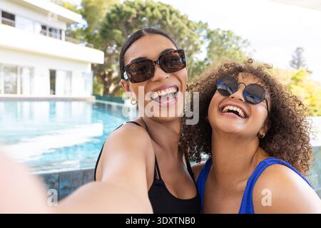 African American female friends posing at modern house pool, taking selfie in shades and swimsuits Stock Photo