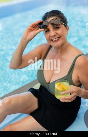 Female smiling and lifting dark sunglasses holding citrus drink poolside in olive-green swimsuit Stock Photo