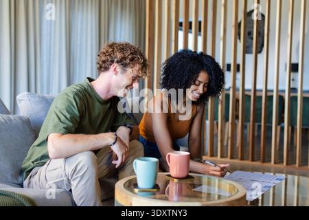 Diverse couple sitting on light-gray sofa in modern living room, reviewing documents with pen Stock Photo