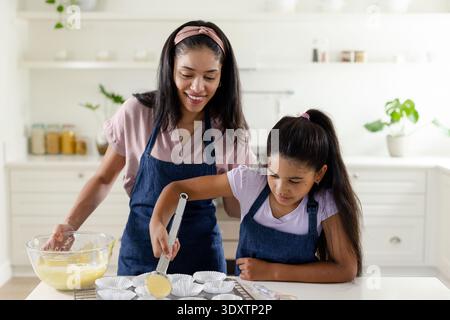African American mom and child scooping batter from glass bowl into paper liners at home counter Stock Photo
