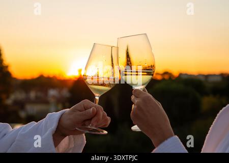 Couple in white bathrobes toasting with glasses of white wine during golden hour. The scene captures a romantic moment on a rooftop with a vibrant sun Stock Photo